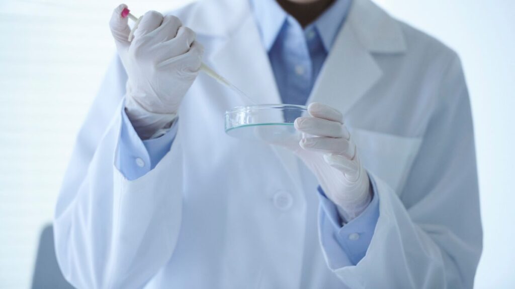 A medical researcher in a white lab coat and gloves holding a pipette over a Petri dish in a laboratory setting.