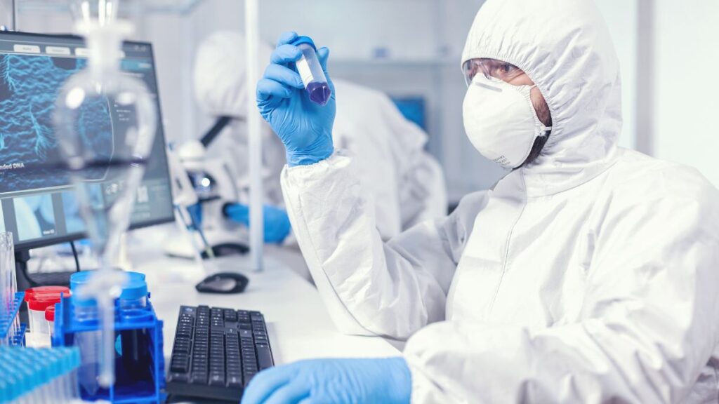 A researcher in a full-body white protective suit, mask, and blue gloves holding up a test tube in a laboratory with scientific equipment and computer screens.