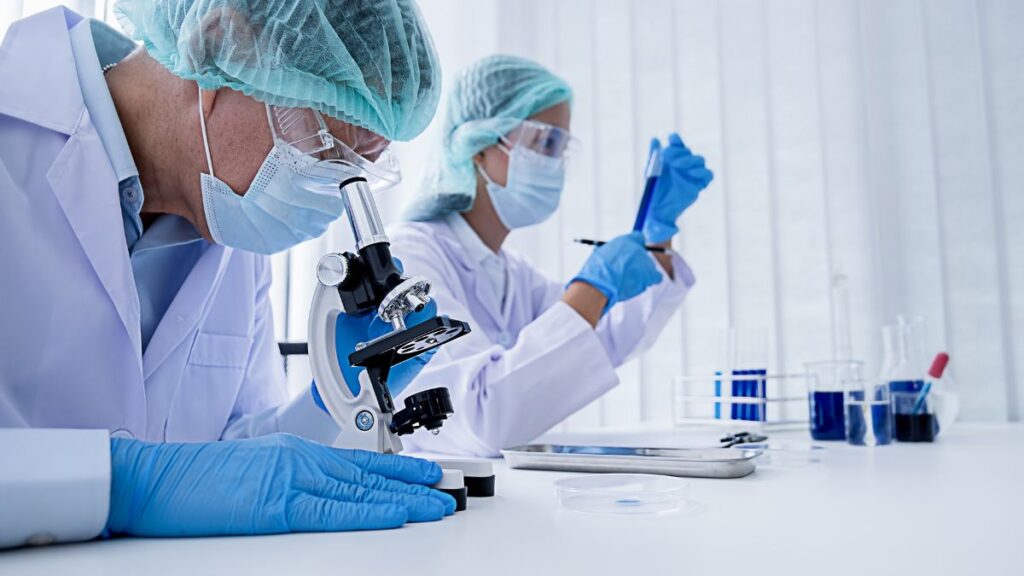 Two researchers in lab coats and protective gear examining samples with a microscope in a bright laboratory.