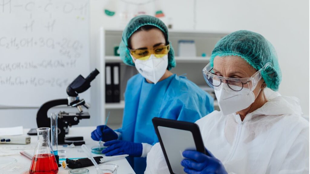 Two researchers wearing protective gear, face masks, and safety goggles reviewing data on a tablet in a laboratory.