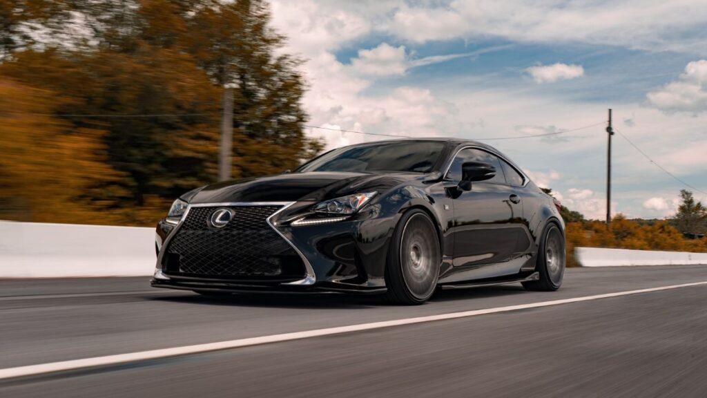 A sleek black Lexus luxury coupe driving on a road surrounded by trees under a cloudy sky.