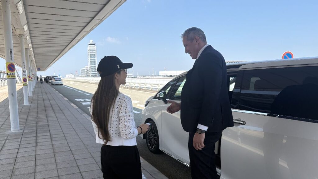 A professional staff member welcoming a client next to a luxury vehicle at Kansai International Airport (KIX).