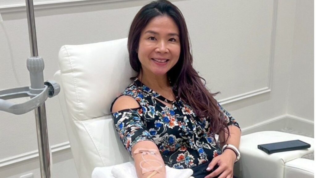 A woman smiling while receiving an IV infusion treatment in a medical room.