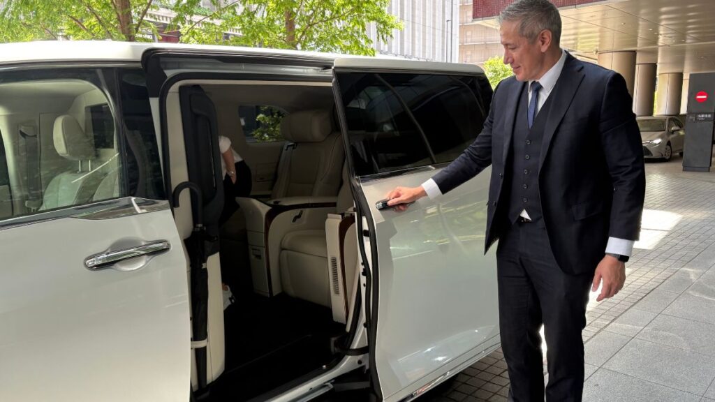 A driver in a suit opening the door of a white Lexus LM minivan for a VIP transfer.